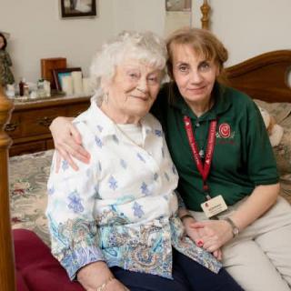 An elderly woman and her caregiver sit together on a bed, sharing a moment of companionship and affection.