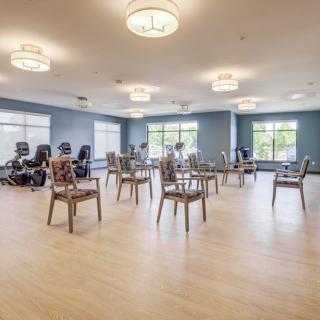 A spacious exercise room with chairs arranged in the center and stationary bikes along the wall, featuring large windows and wooden flooring.