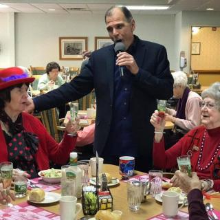 A cheerful gathering of seniors at a dining event, with a man holding a microphone and leading a toast.
