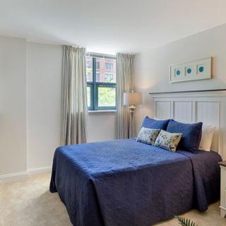 A cozy bedroom with a blue-themed decor, featuring a neatly made bed, artwork on the wall, and natural light from the window.