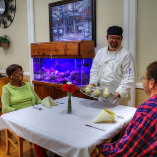 A chef serves a meal to two senior diners in a cozy dining room with an aquarium.