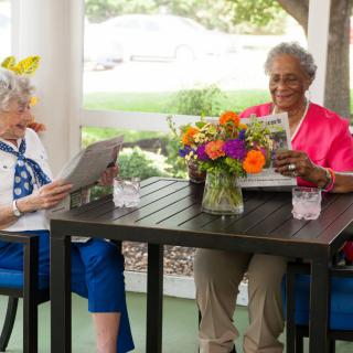 Two senior women enjoying a relaxed afternoon reading newspapers outdoors at a table with a colorful flower arrangement.