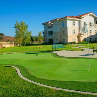 An outdoor putting green with a residential building in the background, surrounded by well-maintained grass and trees.
