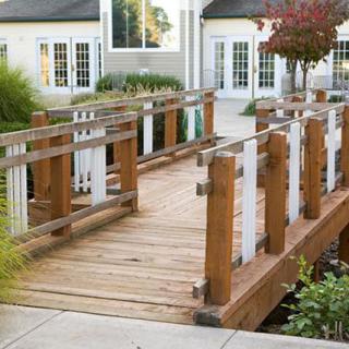A small wooden bridge with sturdy railings leading to a building entrance, surrounded by greenery and landscaped gardens.