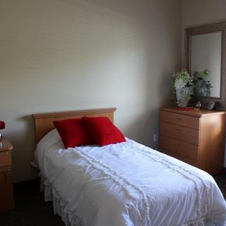 A minimalist bedroom with a single bed adorned with red pillows and white bedding, alongside a dresser and mirror.