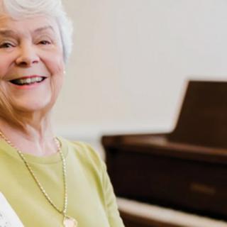 An elderly woman smiling warmly in front of a piano.