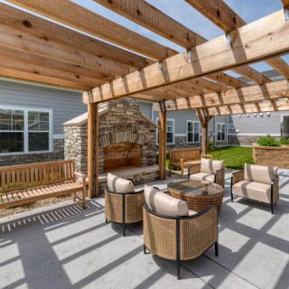 A cozy outdoor seating area under a wooden pergola, featuring comfortable chairs and a stone fireplace.