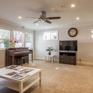 A cozy living room with a piano, television, and coffee table, featuring a ceiling fan and natural light from multiple windows.