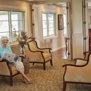 An elderly woman reads a newspaper in a comfortable lounge area.