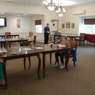 A dining room with several seniors seated at tables, featuring a chandelier and large windows.