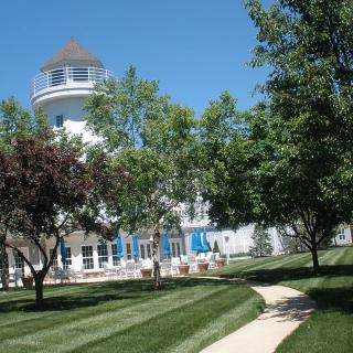 A scenic view of a building resembling a lighthouse, surrounded by trees and a well-maintained lawn under a clear blue sky.