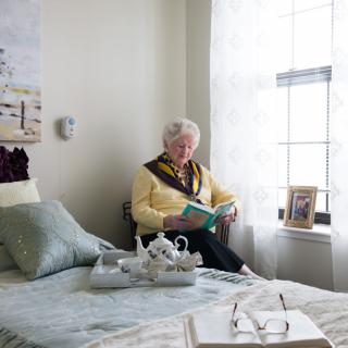 An elderly person sitting by the window, reading a book in a cozy bedroom with a tea set on the bed.