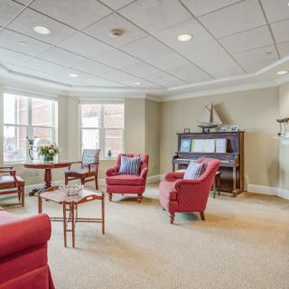 A cozy living room with red chairs and a piano, featuring large windows allowing natural light.