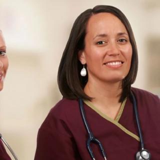 Two nurses in uniform with stethoscopes, smiling in a healthcare setting.
