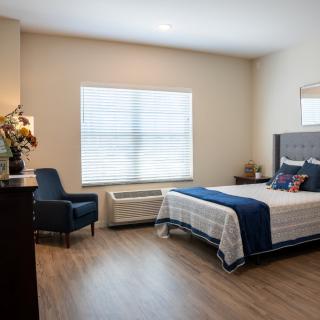 A modern bedroom with minimalist decor, featuring a bed, chair, and a large window providing natural light.