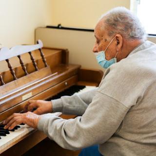 An elderly man playing piano indoors while wearing a mask.