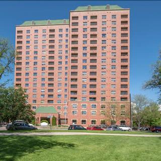 A tall residential apartment building with multiple floors, surrounded by trees and parked cars under a clear blue sky.