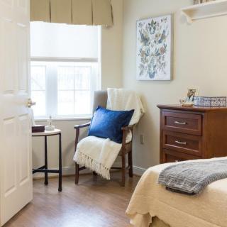 A cozy bedroom with a chair, pillow, and blanket next to a window letting in natural sunlight.