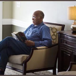 A man sitting in a chair reading a book beside a lamp.