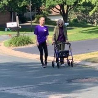 A caregiver assisting an elderly person using a walker on a sunny street.