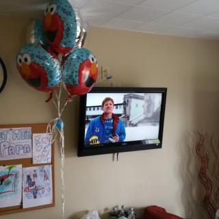 A living room featuring balloons and a television playing a show, with children's artwork displayed on a corkboard.