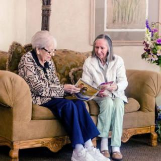 Two elderly women sit on a couch together, engaged in reading and conversation.
