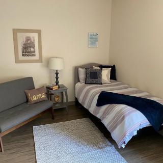 A minimalist bedroom featuring a single bed with striped bedding, a small sofa, a table lamp, and simple wall decor.