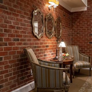 A cozy seating area with armchairs and a table against a brick wall, adorned with mirrors and illuminated by a lamp.