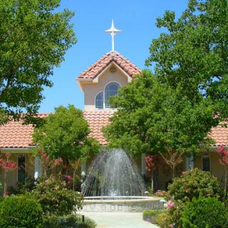 A serene church with a fountain, surrounded by lush greenery and a clear blue sky.