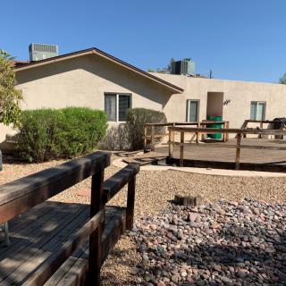 A single-story house with a wooden ramp, surrounded by desert landscaping and gravel.