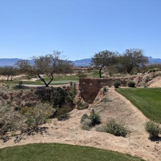 A scenic view of a golf course in a desert setting, featuring green grass, trees, and mountains in the background.