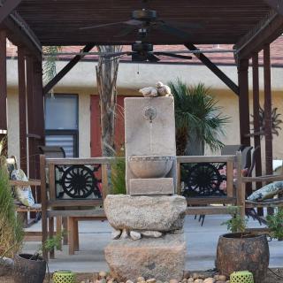 A stone fountain under a wooden gazebo surrounded by plants in an outdoor setting.
