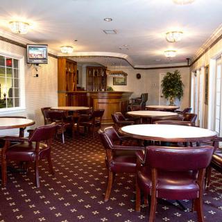 A cozy dining area with round tables and upholstered chairs, featuring carpeted flooring and large windows.
