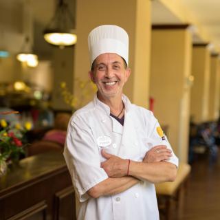 A chef in uniform standing in a kitchen setting, smiling confidently.