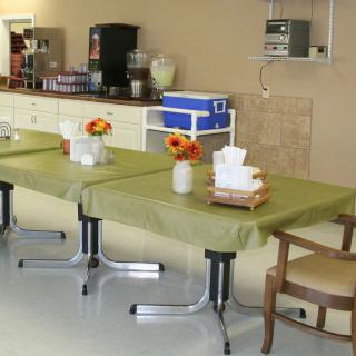 A room with tables covered in green tablecloths and decorated with flower arrangements, alongside a kitchen area.