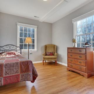 A cozy bedroom featuring a single bed, armchair, dresser, and ample natural light from two windows.