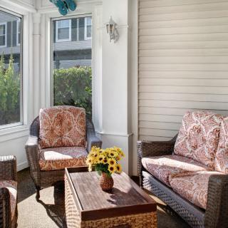 A cozy enclosed porch with wicker furniture, featuring armchairs and a sofa adorned with patterned cushions. A wooden table with a vase of sunflowers adds a cheerful touch.