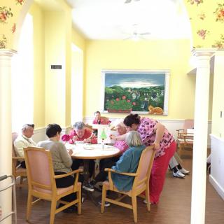 A group of elderly individuals seated around a table in a brightly lit room, engaged in conversation or activities.