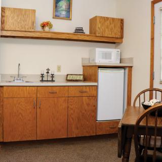 A compact kitchenette with wooden cabinets, a microwave, and a small refrigerator next to a dining table with chairs.