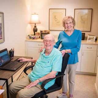 An elderly couple smiling in their home office, with the man seated at a desk using a laptop.