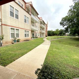 A sidewalk leads to a multi-story apartment building surrounded by a well-maintained lawn and trees on a cloudy day.