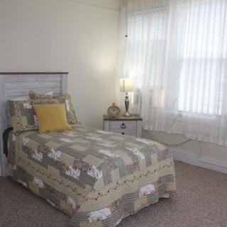 A cozy bedroom with a single bed, decorated with patterned bedding and a yellow accent pillow. A lamp sits on the nightstand beside sheer curtained windows.