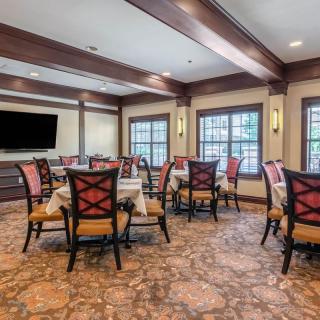 A cozy dining room with multiple tables and chairs, featuring large windows and a television on the wall.