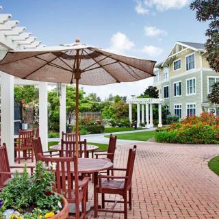 A charming outdoor patio with tables and chairs under large umbrellas, surrounded by a beautifully landscaped garden and adjacent to a multi-story building.