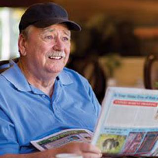 A man in a hat is smiling while reading a newspaper indoors.