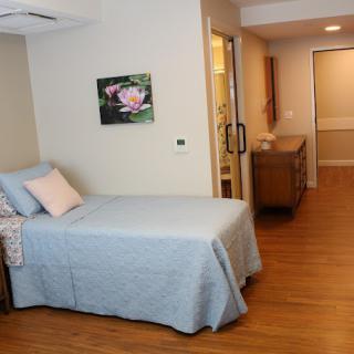A clean and minimalist hospital room featuring a single bed with a light blue cover and wooden flooring.