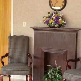 A waiting room with two chairs, a mantelpiece decorated with flowers, and a wall clock above.