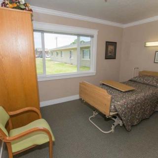 A simple nursing home room with a single bed, wooden wardrobe, and a large window providing natural light.