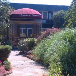 A building entrance with a maroon canopy, surrounded by lush greenery and a stone pathway.