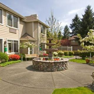 A peaceful courtyard with a central stone fountain surrounded by lush greenery and flowers, adjacent to a residential building.